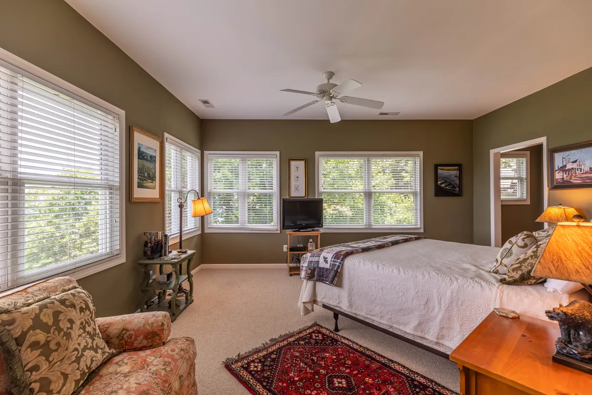 A cozy bedroom with a comfortable bed, armchair by the window, soft lighting from a bedside lamp, and a television. The windows let in natural light and possibly offer views of the surrounding landscape.