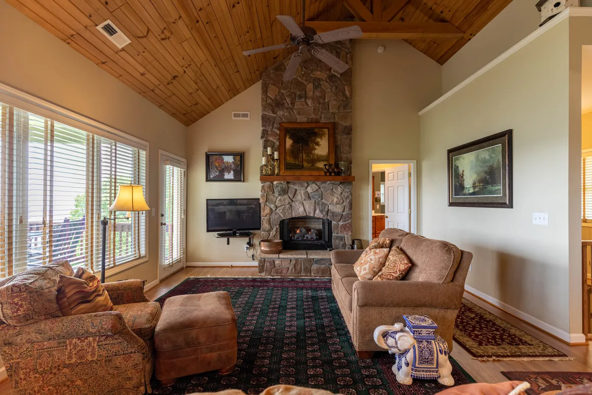 A warm, inviting living room with a prominent stone gas-log fireplace, wooden ceiling, comfortable seating, and artwork on the walls. The large windows allow natural light to bathe the space, creating a cozy and relaxing atmosphere.