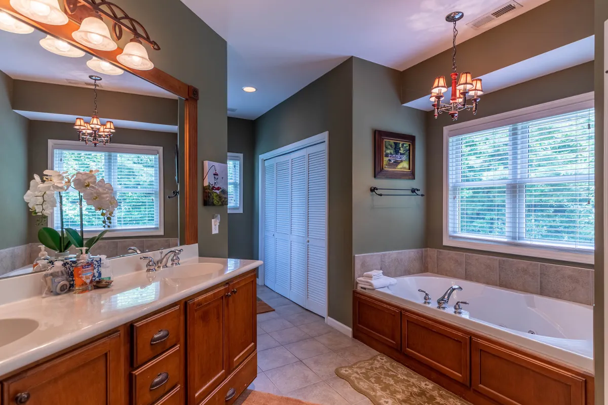 A serene bathroom featuring a double vanity, spacious tub, a window with views of greenery, and a decorative chandelier. The space is airy, luxurious, and designed for relaxation.