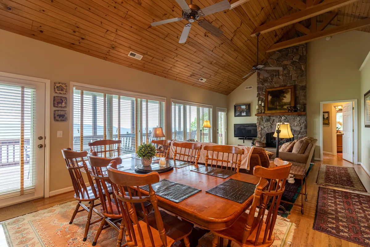 A cozy dining area centered around a stone fireplace with large windows that provide wide views of the surrounding landscape. The wooden ceiling, warm lighting, and inviting setup set a scene perfect for both summer evenings and winter gatherings, reflecting comfort after a day outdoors.