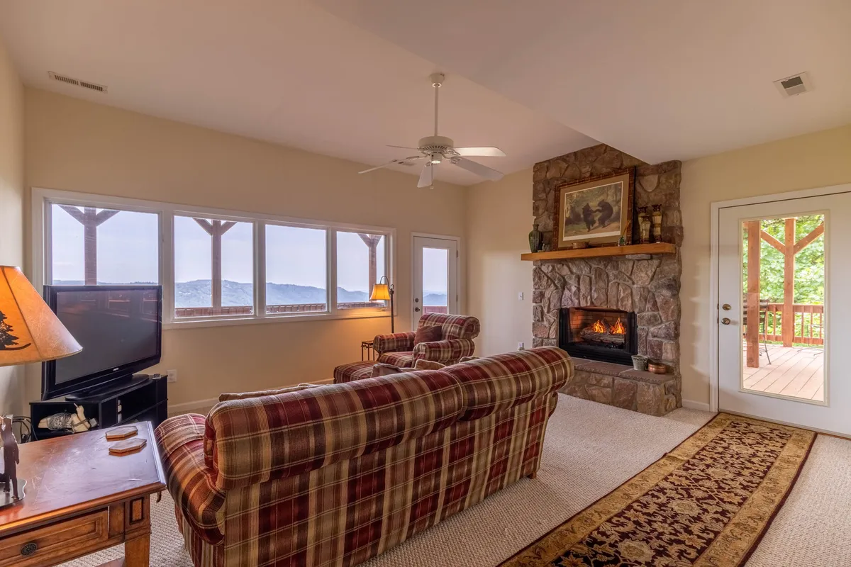 A living area with a warm fireplace, plaid sofas, large windows offering picturesque views, and a wooden door leading outside. The space is bathed in natural light, inviting both relaxation and social connection.