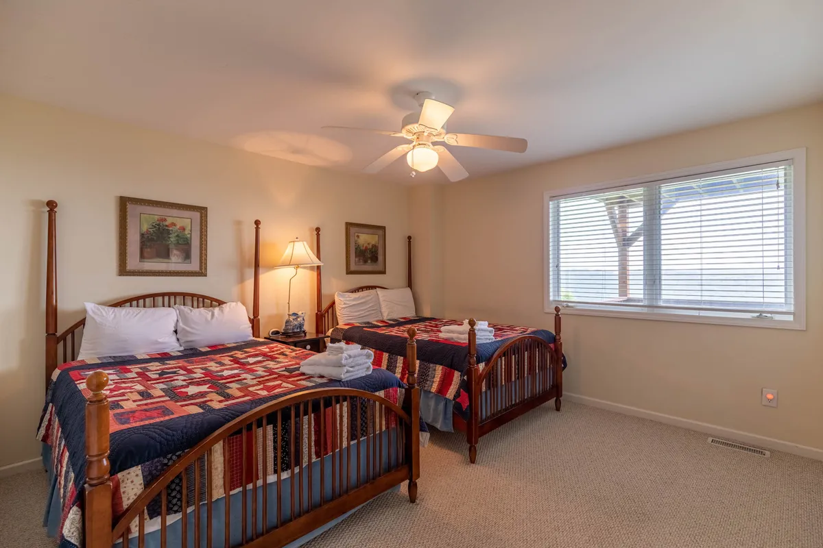 A private sleeping area with two wooden beds, both topped with cozy quilts, a table lamp between, and a window with blinds. The simple, inviting setting is perfectly suited for kids, teens, or extra guests.