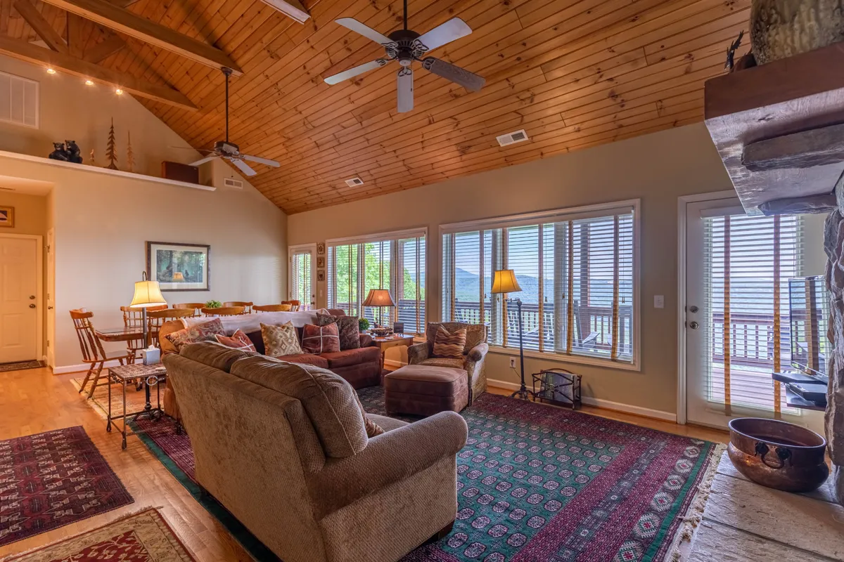 A welcoming open-concept living area featuring comfortable sofas, a grand stone fireplace, large windows that frame mountain views, and an adjacent dining area. The wooden vaulted ceiling adds spaciousness and warmth to the room.