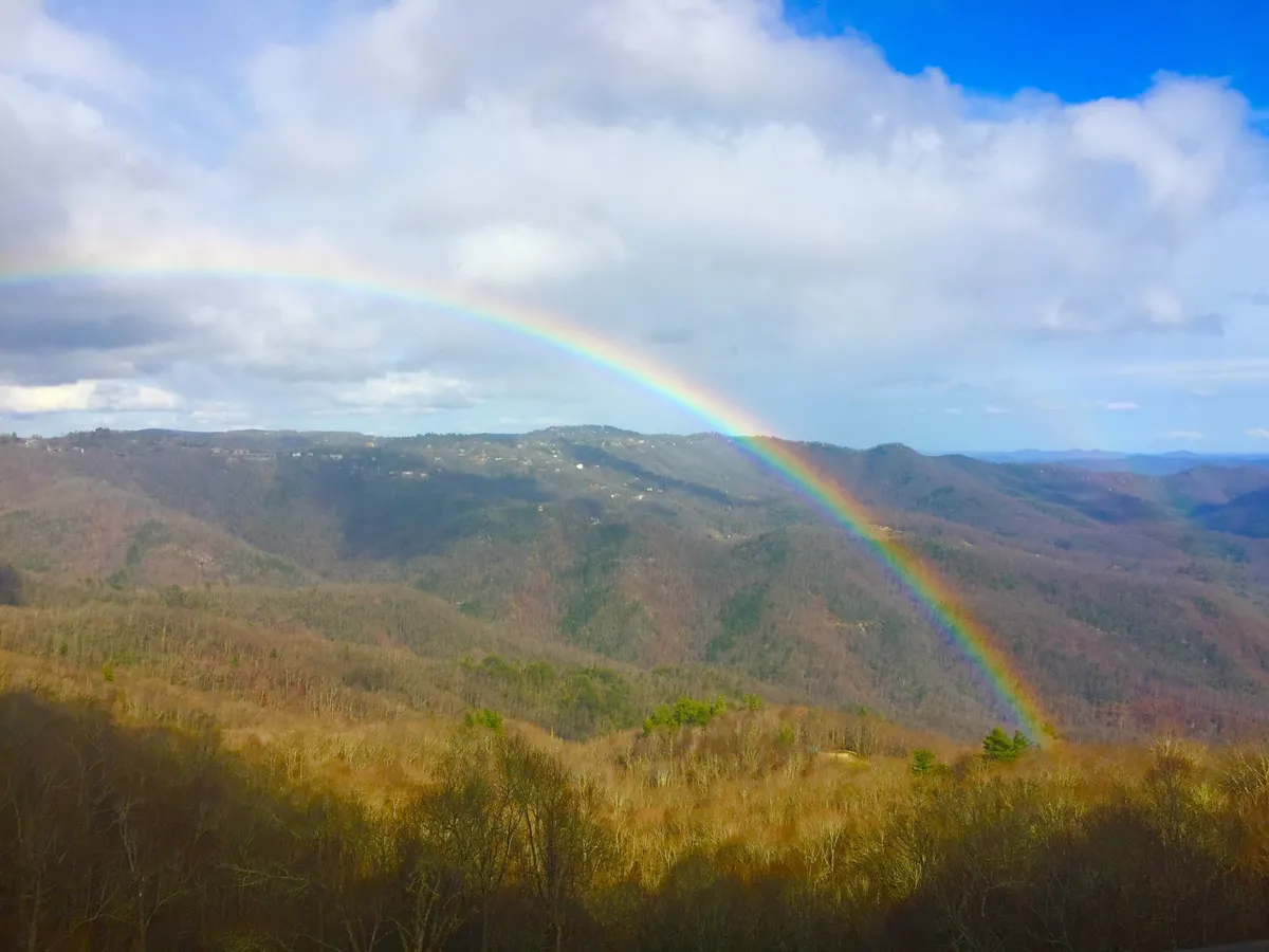 A vibrant rainbow arches over a sweeping mountain landscape under dramatic clouds, creating a postcard-worthy scene. The image communicates awe-inspiring natural beauty, ideal for hiking, photography, and outdoor adventures.