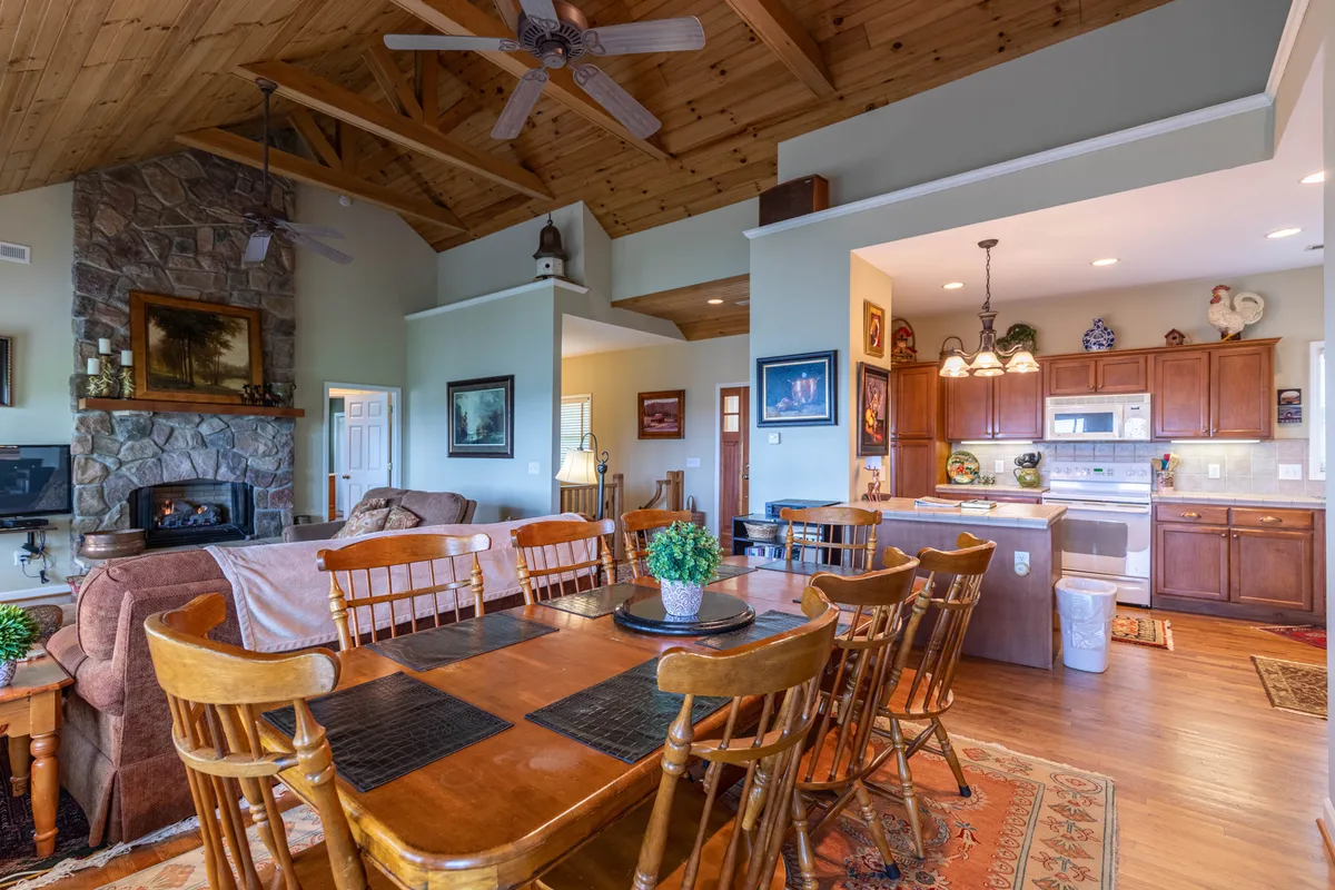 A broad view of the main level’s heart: a stone fireplace anchoring the living area, a spacious dining table nearby, wooden ceiling beams above, and a glimpse of the connected kitchen. The setting feels inviting and perfect for family gatherings.